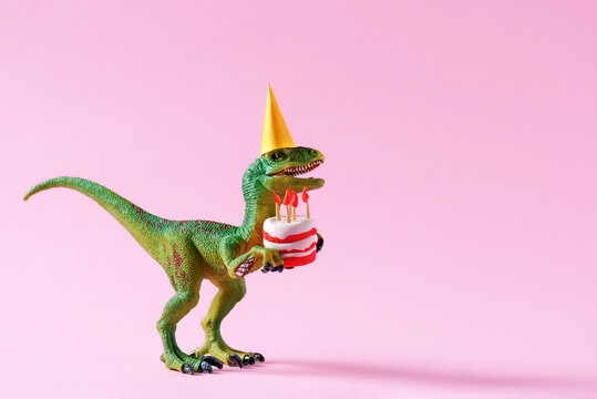 Fototapeta Cute happy green dinosaur in birthday hat holding cake with flaming candles on pastel pink background. Copy space. Minimal art birthday card idea.