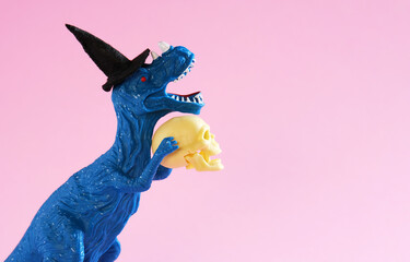 Happy blue dinosaur in witch hat holding human skull on pink background. Minimal humor card for...