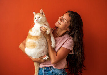 Happy young brunette caucasian woman holding her white and red cat on orange background.
