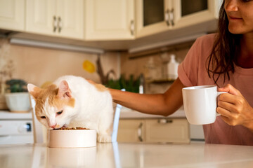 Beautiful young woman drinking coffee and feeding cat, sitting at kitchen table.