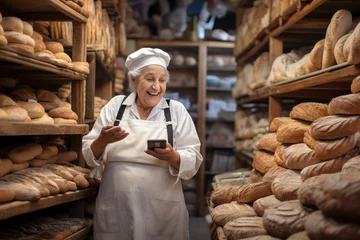 Foto op Plexiglas A old woman seller in bakery store with hold phone and in smile  © MAXXIMA Graphica