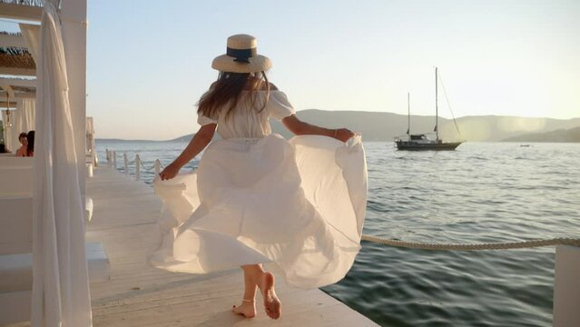 Slow-motion of young brunette in white dress, hat, and flowing hair runs on wooden pier by sunset at sea. Evokes tourism, summertime, holiday, and beach vacation.