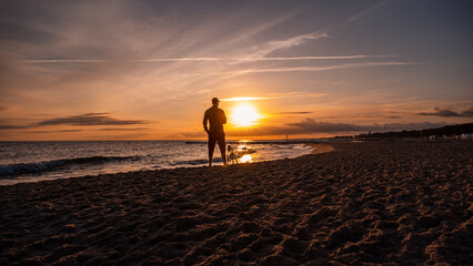 Fototapeta na wymiar A man standing in front of the sunrise at the Baltic Sea. Next to the man is a camera on a tripod. Sandy beach in Kolobřeh. Kołobrzeg is city in Poland.