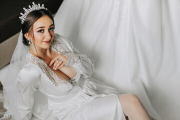 An incredibly beautiful bride in the morning in a satin robe with a tiara on her head poses next to...