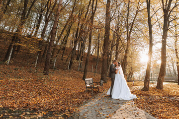 A fabulous romantic newlywed couple is hugging at sunset in an autumn park with leaves from trees on the grass. Wide angle photo