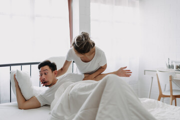 Funny asian couple wife try to wake lazy husband up in white bedroom.
