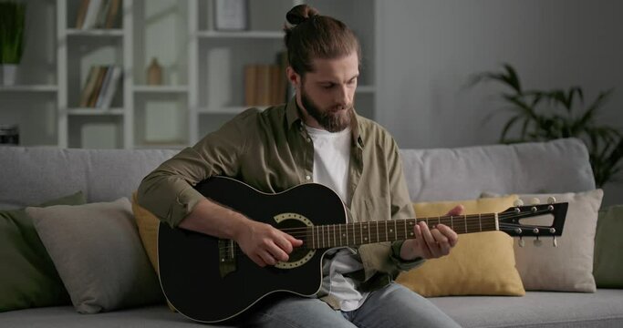 Attractive man with long hair sitting on sofa and playing acoustic guitar.