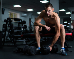 Muscular shirtless man sitting on a bench and lifting a dumbbell. 