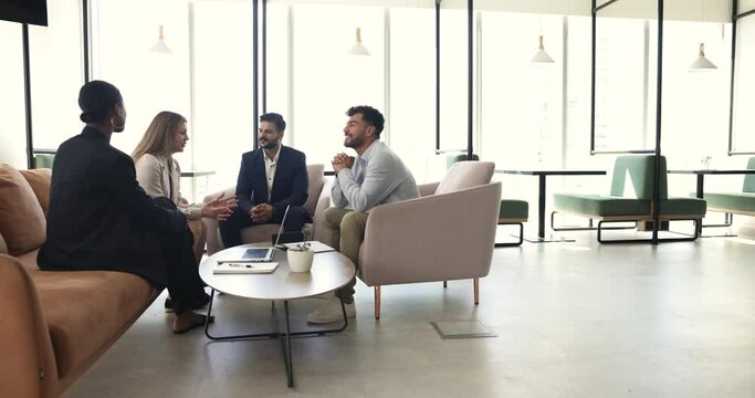 Indian businessman and diverse office employees, friendly teammates talking seated on armchairs in modern co-working space. Teamwork and briefing event of four professional staff members, leadership