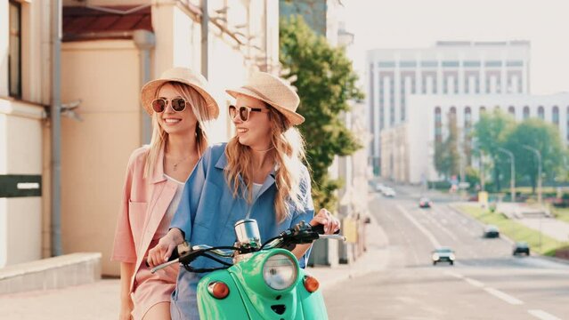 Two young beautiful smiling hipster female in trendy clothes. Carefree women driving retro motorbike in the street. Positive models having fun, riding classic Italian scooter in eyewear and hat