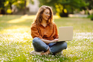 Portrait of a young woman sitting on the grass in the park working, studying on a laptop. Happy...