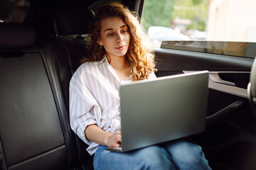 Stylish woman with glasses in the car, sitting on the back seat of the car and working on a laptop. Business concept, blogging, freelancing.