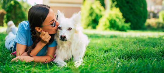 Happy woman hugging and kissing her white fluffy dog on the outdoor in the park on lawn. Adoption, rescued, shelter, companion, pet, best friend.