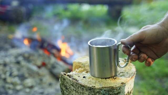 A man takes a mug of hot herbal tea from a stump near the fire. The sharpness moves from a mug of tea to a fire.