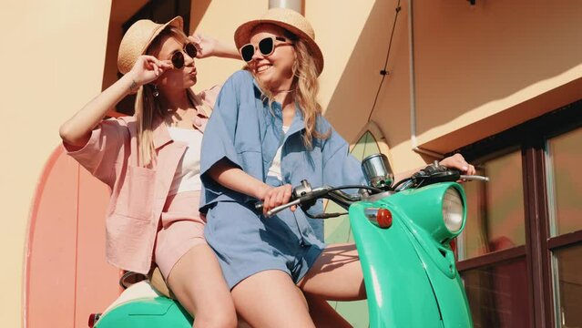 Two young beautiful smiling hipster female in trendy clothes. Carefree women driving retro motorbike in the street. Positive models having fun, riding classic Italian scooter in eyewear and hat