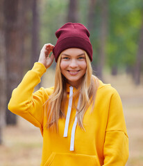 Attractive blue eyed blonde woman walk on the park. Girl wear yellow hoodie and burgundy hat. Portrait of a joyful young woman enjoying in autumn park.