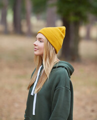 Attractive blue eyed blonde woman walk on the park. Girl wear green hoodie and yellow hat. Portrait of a joyful young woman enjoying in autumn park.