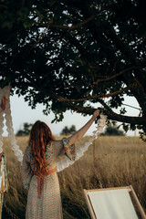 red-haired young woman in a dress pulls a paper garland on a tree. woman with wavy hair decorates outdoors party place.