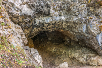 Limestone cliff wall with entrance of the cave. Cave in a layered limestone rock. - obrazy, fototapety, plakaty