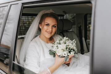 A beautiful smiling bride on her wedding day is sitting in a car and holding a wedding bouquet of white and pink roses. A young woman with a beautiful hairstyle rejoices on her wedding day