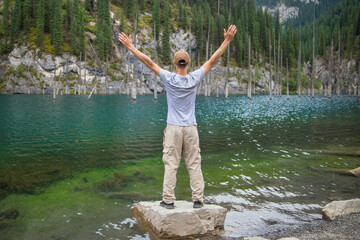 Tourist man travels through the mountains of Central Asia, standing with arms outstretched and looking at the mountain lake Kaindy with sunken fir trees.