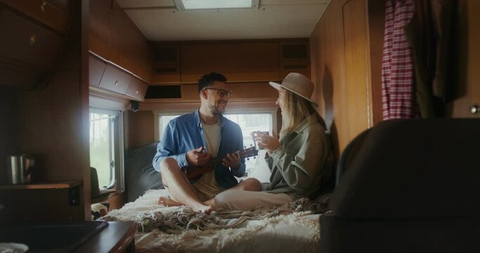 A man plays the ukulele and sings looking at the woman sitting next to him on the bed. A woman holds a mug with a hot drink in her hands. Romantic van ride