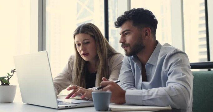 Two multiethnic office employees, coworkers working together on laptop, look at device screen makes on-line project corrections, engaged in teamwork. Mentor and apprentice using computer at workplace