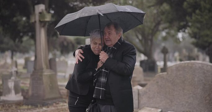Support, death or old couple in cemetery for funeral, service or burial for repsect in Christian religion. Umbrella, depressed or sad senior people hug for comfort in graveyard crying in mourning