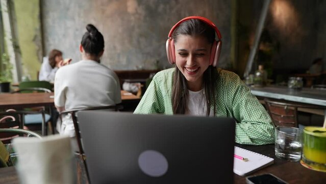 A young girl wearing headphones is having a conversation on her laptop in a café.
