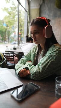 A café backdrop presents a young and pretty girl using a laptop with headphones on.