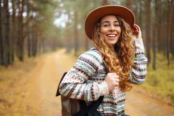 Active young woman with a backpack and a hat enjoys a beautiful autumn day in the forest, exploring the thicket of the forest. Traveler is walking along a footpath in the forest.
