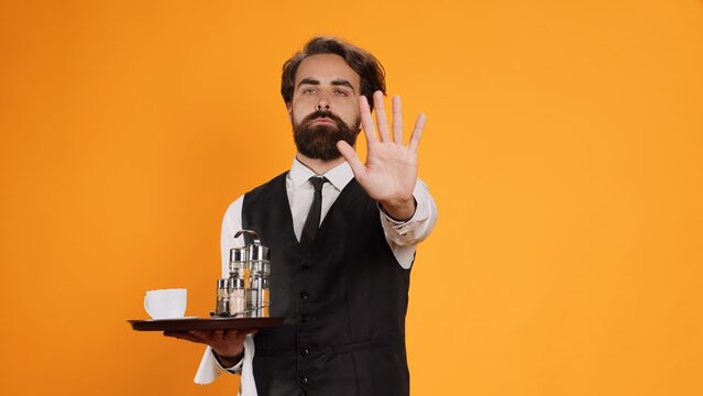 Expert valet posing with meal tray and showcasing stop signal with his palm. Staff of luxury restaurant becoming dissatisfied and rejecting other ideas, declining to comply with them.