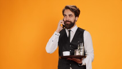 Eatery waiter making dinner appointments over phone while employed by catering sector over yellow background. Staff member receiving call to book table on smartphone line, serving tray.
