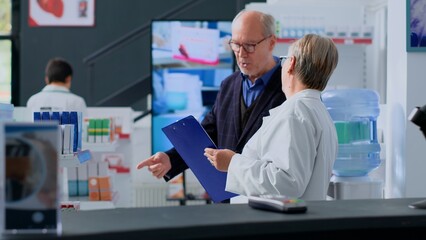 Elderly shopper watching pharmaceutical product advertising on digital screen in apothecary, approaching pharmacist with intention of buying advertised drugs. Expert assists senior customer with info