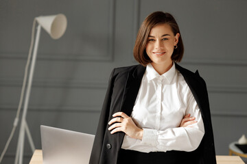 A smiling business woman wearing a white shirt and a black jacket on her shoulders is standing with her arms crossed in front of her on a gray background with a laptop on the table