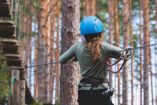 View of high ropes course, kids of climbing in amusement acitivity rope park, passing obstacles and zip line on heights, children teenagers in equipment gear between the trees on heights, summer day - Powered by Adobe