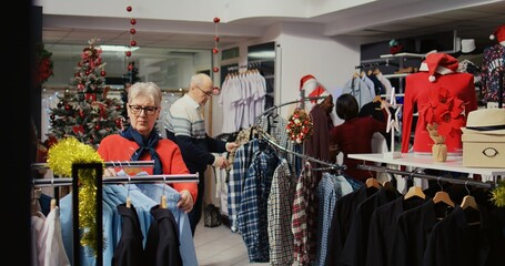Customers roaming around busy clothing store decorated with Christmas ornaments during winter holiday season. Clients shopping for gifts during xmas promotional sales in fashion boutique
