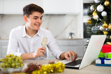 Happy young man with glass of champagne wishes friends merry christmas via internet using laptop