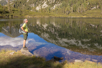 Naklejka premium A man with a backpack stands against the backdrop of a mountain lake with a reflected sky, mountain and clouds