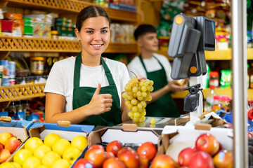 Smiling european female seller weighing fresh grape in supermarket and showing thumbs up