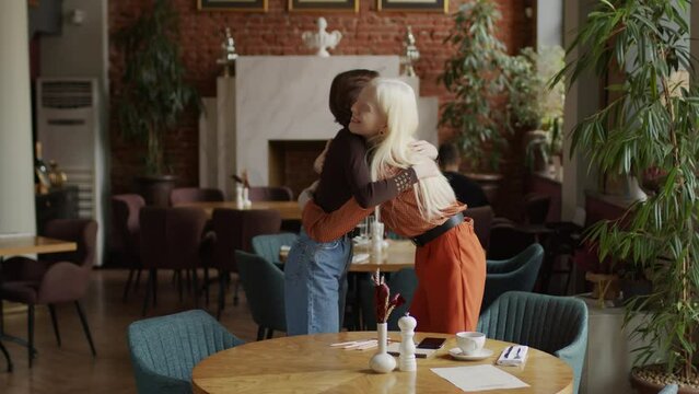 Gen Z albino girl sitting at table in restaurant, seeing her best friend, greeting her with hug then taking seats and chatting