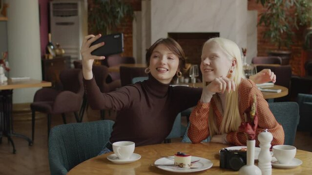 Two cheerful gen Z friends spending time together in modern restaurant taking selfies on smartphone camera
