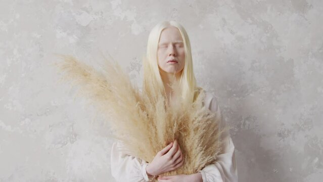 Medium length studio portrait of young albino woman wearing white clothes holding dried bouquet standing with eyes closed against white wallMedium length studio portrait of young albino woman wearing 