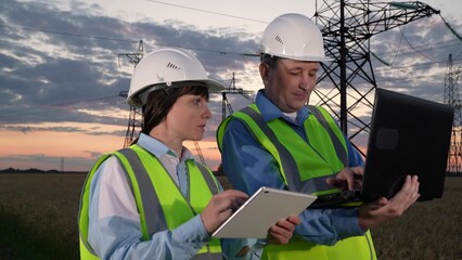 Engineers with colleague study data on computers by power transmission lines