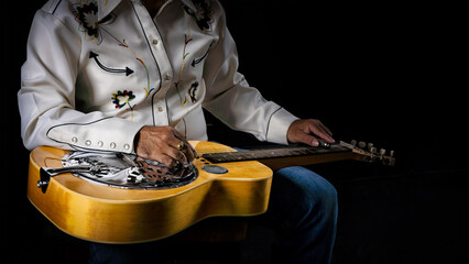 Close-up of a Dobro guitar resting on the lap of a musician dressed in cowboy attire on dark backdrop - obrazy, fototapety, plakaty