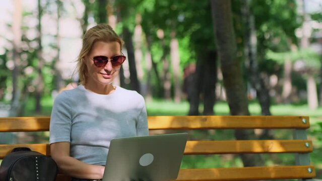 Relaxed blonde woman sitting on the bench using a laptop, close up. Cute female freelancer making remote work in the summer city park on blurred green foliage background.