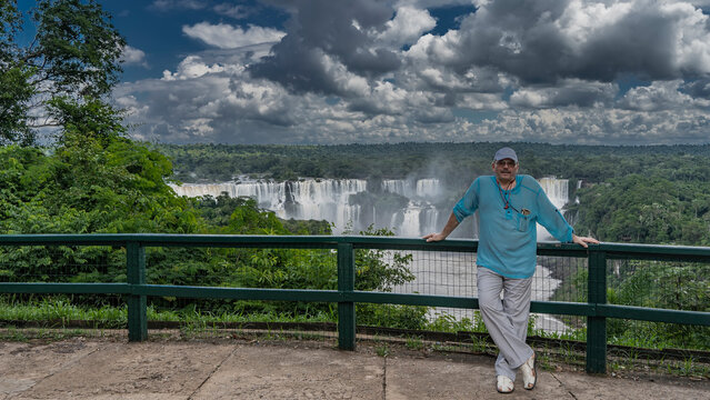 Beautiful waterfall landscape. A man is standing on the observation deck, leaning on the railing. Behind it is a cascade of waterfalls, lush tropical vegetation. Clouds in the sky. Iguazu Falls. Brazi