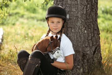 Portrait of a jockey girl in a helmet and a white T-shirt, who sits under a tree with a toy horse...