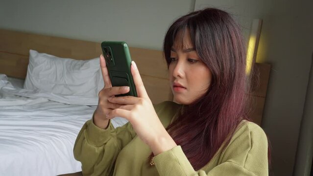 Asian woman sitting in her room while scrolling on her phone
