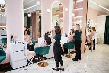Young women sitting in front of mirrors while hairdressers styling clients hair and makeup artist doing professional makeup. Makeup specialist and hairstylists working in modern beauty salon.
